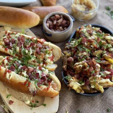 Hot dog în chifle de casă cu varză murată, ceapă caramelizată, bacon crocant, ketchup și maioneză, alături de o tavă rustică de cartofi loaded cu toppinguri. Fundal cu chifle proaspăt coapte, atmosferă casual-rustic