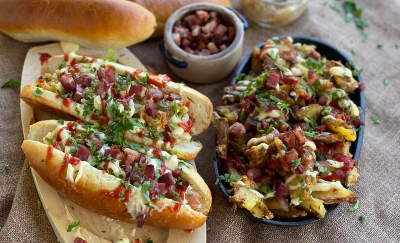 Hot dog în chifle de casă cu varză murată, ceapă caramelizată, bacon crocant, ketchup și maioneză, alături de o tavă rustică de cartofi loaded cu toppinguri. Fundal cu chifle proaspăt coapte, atmosferă casual-rustic