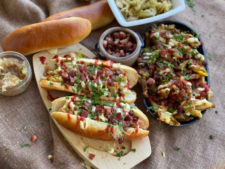 servire-hot-dog-varza-murata-loaded-fries Hot dog servit în chiflă de casă, cu toppinguri de varză murată, ceapă caramelizată, bacon crocant și sosuri, alături de cartofi loaded fries