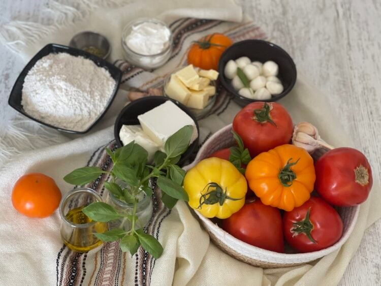 ingrediente-galette-rosii-branza-1 Ingrediente pentru galette: roșii de diferite culori, cremă de brânză, brânză feta, usturoi, ierburi aromate, unt, făină, smântână și ou, așezate pe fundal rustic.