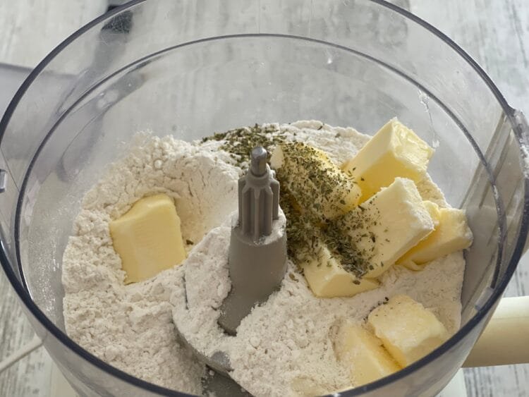 Ingrediente pentru aluatul de galette – făină, unt rece și oregano – în bolul unui robot de bucătărie.
