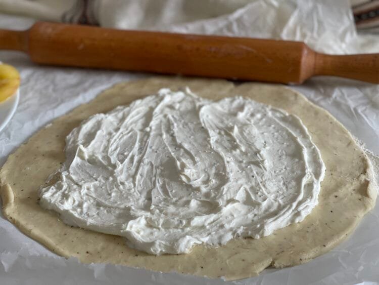 Cremă de brânză întinsă uniform pe aluatul rotund, lăsând margini libere pentru împachetare, în procesul de asamblare a unei galette rustice.