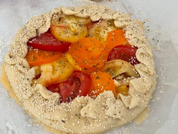 Galette formată, cu marginile împăturite, unsă cu ou și decorată cu semințe de susan, pe hârtie de copt, înainte de coacere.