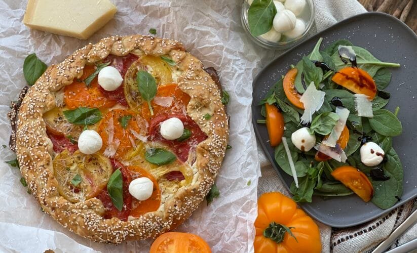 Tartă galette rustică cu roșii colorate și cremă de brânză, decorată cu frunze de busuioc și mini mozzarella, așezată pe hârtie de copt.