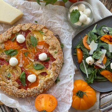 Tartă galette rustică cu roșii colorate și cremă de brânză, decorată cu frunze de busuioc și mini mozzarella, așezată pe hârtie de copt.