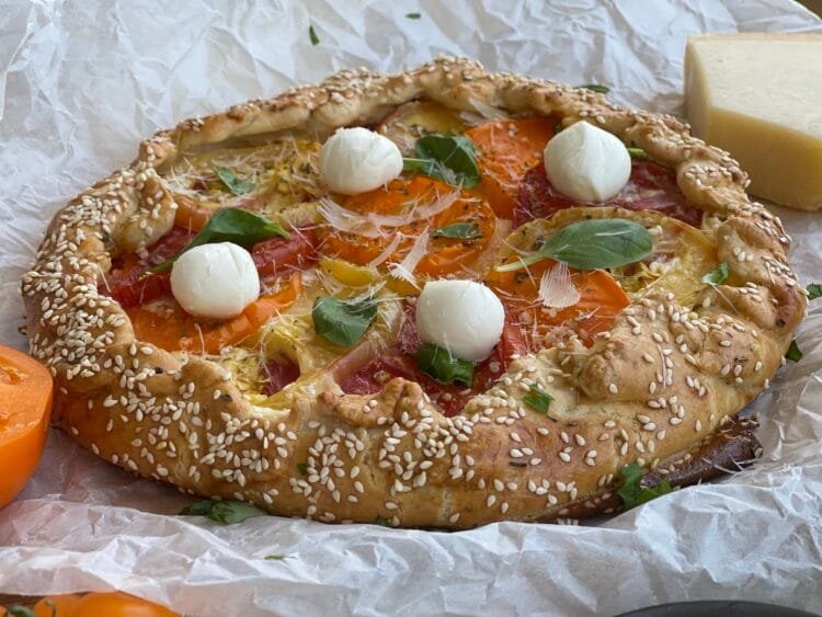 Galette rustică, coaptă perfect, decorată cu mini mozzarella, busuioc proaspăt și fulgi de parmezan, pe hârtie de copt.