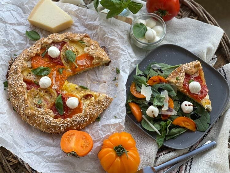 Galette rustică cu roșii, parțial feliată, pe hârtie de copt; în prim-plan, o felie servită pe farfurie alături de salată verde.